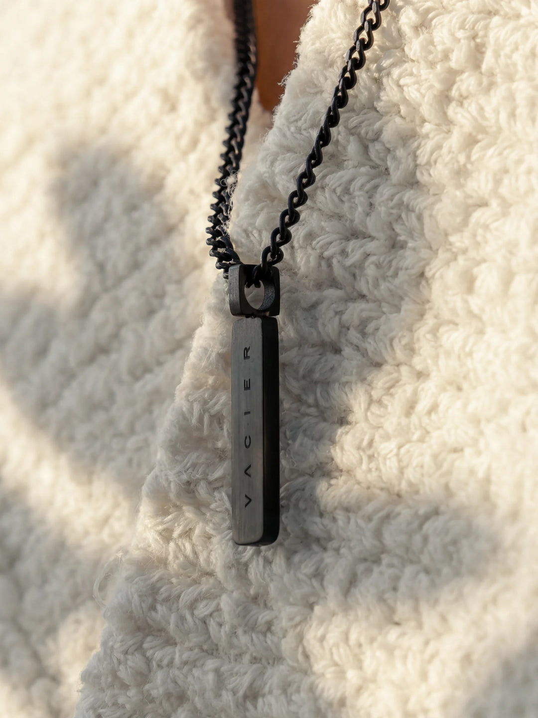 Close-up of a black necklace with a rectangular pendant against a light-colored garment 