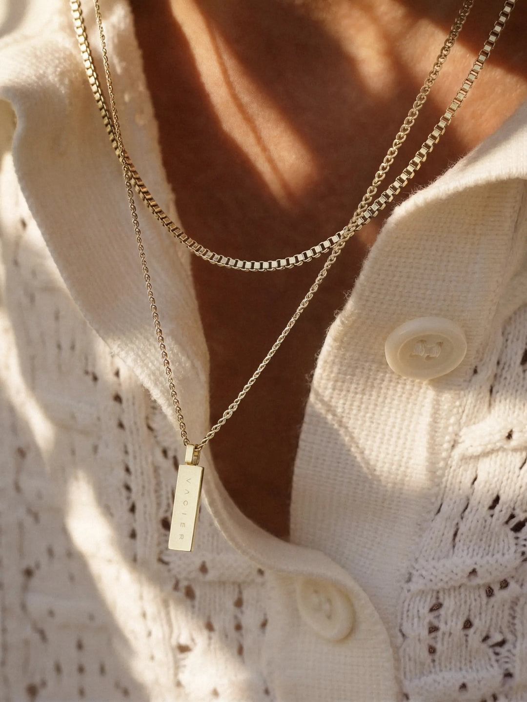 Close-up of a person wearing two gold necklaces, one of which has a rectangular pendant, with a light-colored shirt