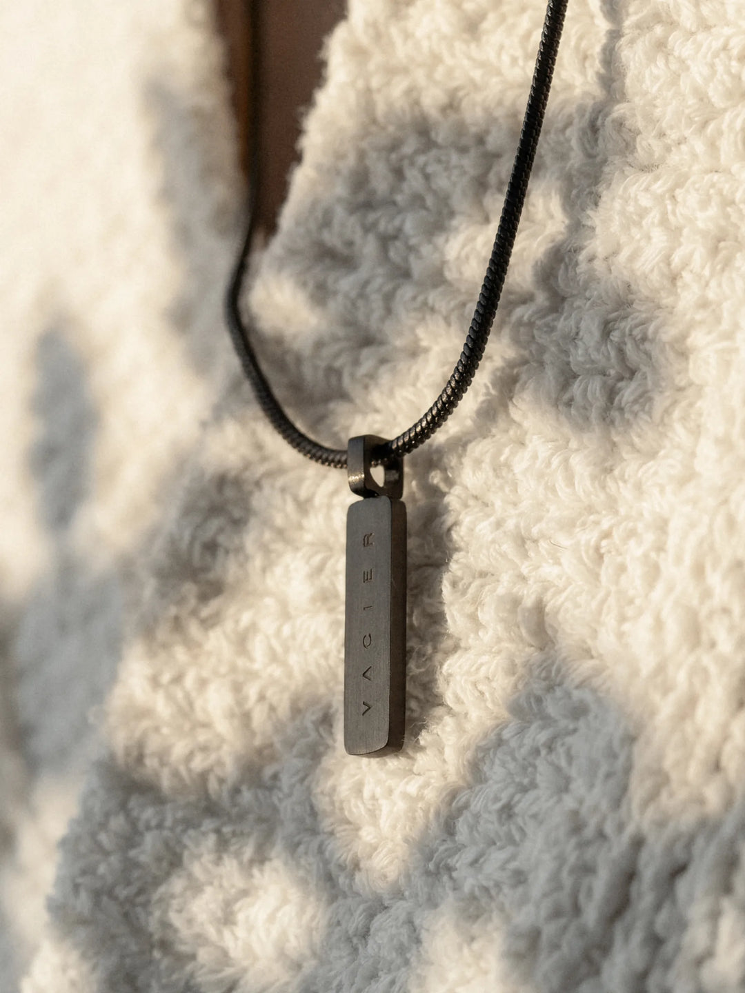 Close-up of a black necklace with a rectangular pendant against a white textured garment