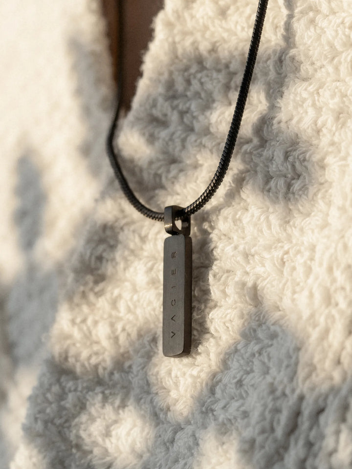 Close-up of a black necklace with a rectangular pendant against a white textured garment