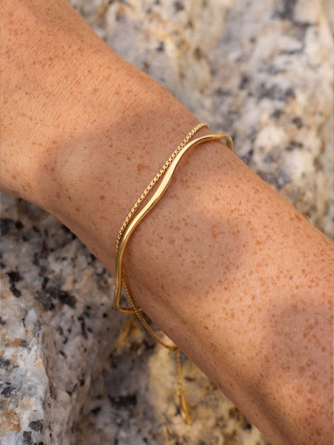 A gold cuff and a gold chain bracelet on a wrist against a textured stone background