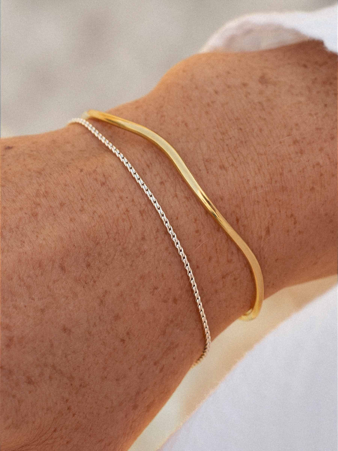 A gold cuff and a silver hain bracelet on a person'e wrist against a light blurred background