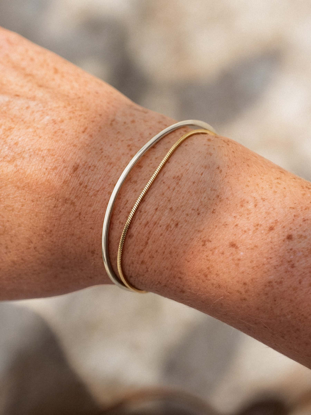 Close-up of a person's arm wearing a silver cuff and a gold chain bracelet with a blurred natural background.