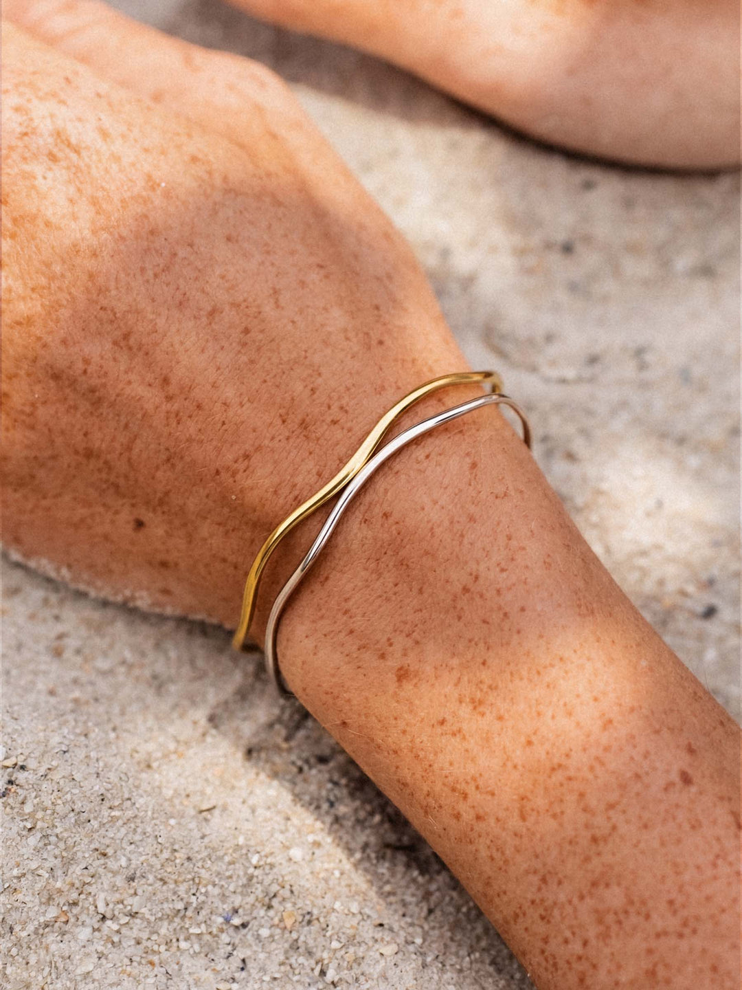 A gold and a silver cuff on a person's wrist against a neutral background