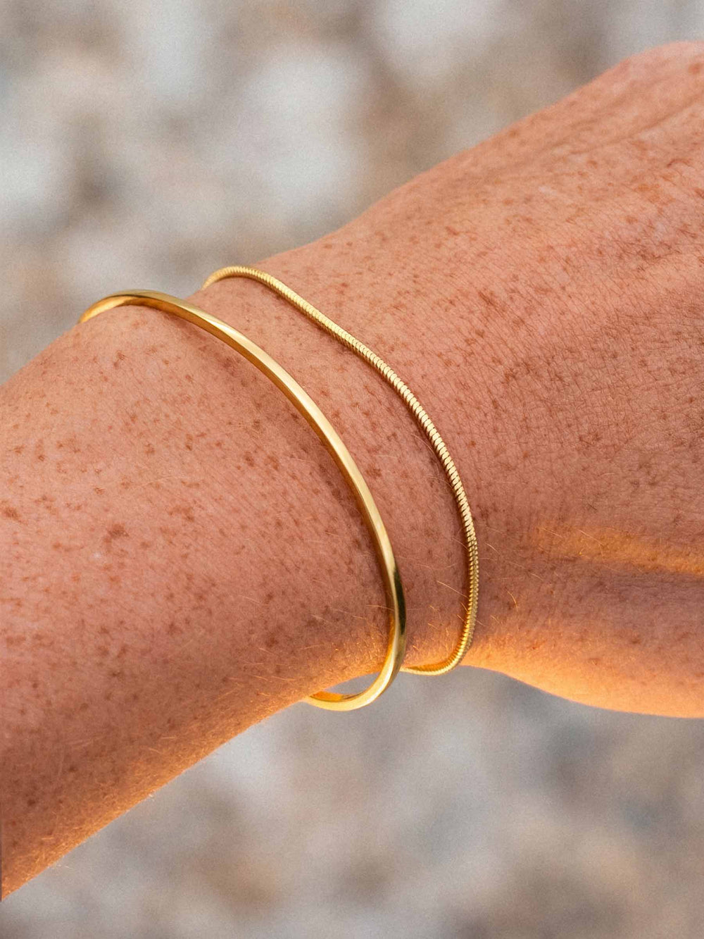 A gold cuff and a gold chain bracelet on a wrist with a blurred natural background