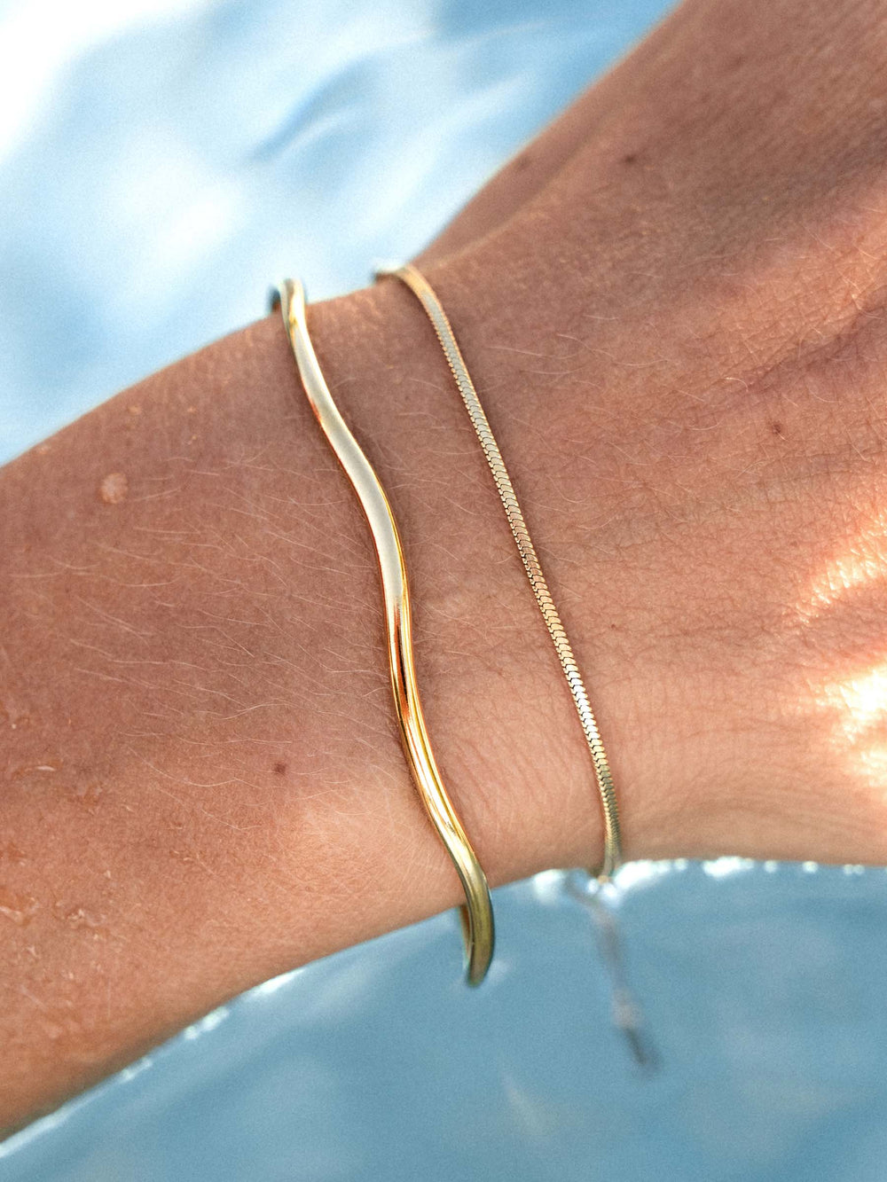 Gold bracelet and a gold cuff on a wrist with a blurred water background